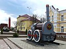 Una locomotora gris, que parece casi de juguete, en medio de una plaza de suelo de adoquines rodeados por tierra. Al fondo, unas casas señoriales amarillas