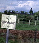 Behind a faded white sign saying "HALT ZONENGRENZE", there is a derelict barbed-wire fence supported by leaning wooden posts; some distance behind that, there are two newer-looking barbed-wire fences supported by concrete posts. Another fence and a guard tower are visible in the distance.