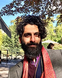Photo depicting Emad Kiyaei, with a black beard and dark hair, smiling enigmatically in afternoon sunlight in a square in Paris.