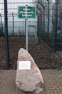 Großer Stein mit eingearbeiteter Metalltafel vor einem Schild mit der Aufschrift „Günter-Schwannecke-Spielplatz“. Dahinter befindet sich ein abgezäuntes Fußballfeld.