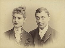  A charming and iconic period photo of a young couple around 1900. The lady has her hair swept upwards and is wearing a high collar fastened with a jewel. The gentleman sports a gallant moustache neatly trimmed and is also wearing a high collar.