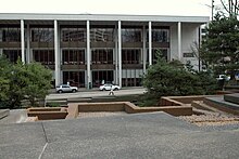Concrete geometric shapes form a platform in the forefront along with a brown border. In the background are trees and two cars parked in front of a rectangular light-colored building with dark windows.