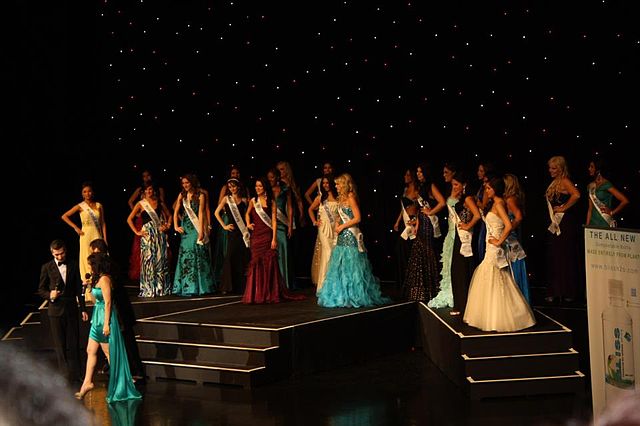 A photograph of a large group of women all wearing differently coloured dresses and standing on a stage in front of a black background