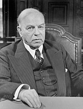 An older man in a three piece suit, seated and looking at the camera