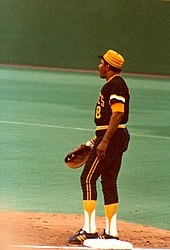 A man wearing a black and gold baseball uniform and baseball glove stands on first base.