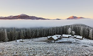 Une colline neigeuse bordée d'une épaisse forêt, chapeautée d'un nuage rasant. Sur la colline une clairière avec quelques chalets.