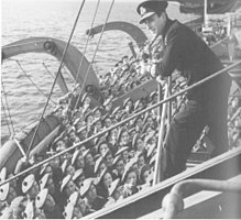 A man in a naval uniform addresses soldiers from a platform on a ship