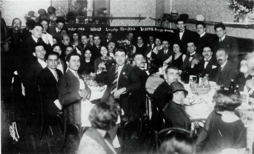 Photographie en noir et blanc de l'intérieur d'un restaurant rempli de convives assis et debouts autour de deux tables.