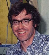 Das Protraifoto eines mittelalten Mannes mit mittellangen, dunklen Haaren. Er trägt eine Brille und lächelt in die Kamera. Weiterhin trägt er ein blaues, wildgemustertes Hemd.