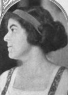 A young woman in profile, with light skin and dark hair in an updo fastened with a wide headband, wearing a light blouse with a square neckline