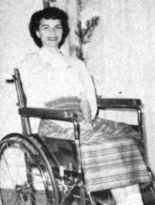 A young white woman with dark hair, seated in a wheelchair, smiling; she is wearing a striped skirt and a white blouse