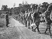 Photographie en noir et blanc de plusieurs militaires japonais marchant les uns derrières les autres sur un chemin en terre et sous la surveillance d'un militaire malaisien.