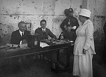Julieta Lanteri, vestida de blanco frente a la mesa de votación en la que está la urna y cuatro hombres observándola.