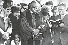 Black and white photo of mourners at a funeral service. Many mourners have their head downs and are visibly dejected.
