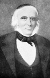 Formal portrait showing the head and shoulders of a white-haired man wearing a dark cape or coat