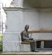 Photographie couleur d'une statue en bronze d'un homme assis sur banc sous un abri en pierre
