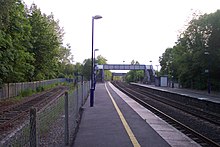 Photograph of a railway station platform, showing the main two pairs of rails, a footbridge, and an old rusted siding