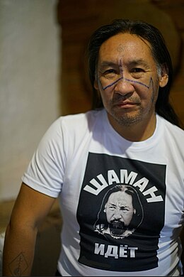 Photographie d'un homme aux cheveux longs portant des tatouages bleus sur le visage et un T-shirt blanc avec son propre portrait.