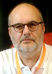 Photographie en buste d'un homme chauve portant une chemise blanche, des lunettes noires et une légère barbe grise.