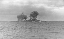 Black-and-white photograph of a warship firing the guns. Two columns of dark smoke drift from the ship