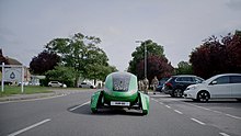 The green, futuristic-looking Kar-go autonomous delivery vehicle driving on the roads through RAF Brize Norton air base during trials in 2021. RAF personnel are walking alongside it and cars are parked beside the road