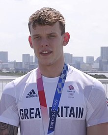 Farbfoto mit Richards unter freiem Himmel im weißen T-Shirt mit der Aufschrift Great Britain. Um den Hals trägt er ein rotblaues Band mit der Aufschrift Tokyo 2020, an dem seine Goldmedaille befestigt ist, die aber in diesem Bildausschnitt nicht zu sehen ist.