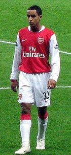 A young black man stands on a pitch dressed in football kit. The body of his shirt is red and bears the club badge and logos of manufacturer and sponsor on the front. The rest of his kit, long sleeves, shorts, socks and boots, is white. His shorts bear the number 32.