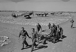 a black and white photograph of men pulling a trolley, with aircraft dispersed in the near background
