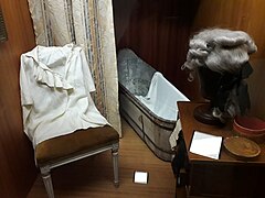 Photographie d'une vitrine du musée. Sur la gauche, sur une chaise, est exposée une chemise française du XVIIIe siècle à col échancré orné de dentelles. À droite, sur une petite commode, une perruque grise de la même époque est disposée sur son support. Au fond, on peut voir une baignoire en fer blanc à demi masquée par un rideau clair.