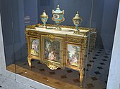 Louis XVI style commode of Madame du Barry; 1772; oak frame, veneer of pearwood, rosewood and kingwood, soft-paste Sèvres porcelain, gilded bronze, white marble, and glass; height: 0.87 m, width: 1.19 m, depth: 0.48 m[98]