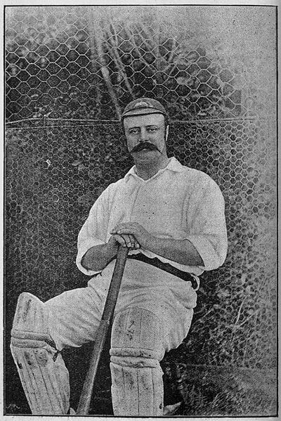 Man with moustache seated on chair holding cricket bat.