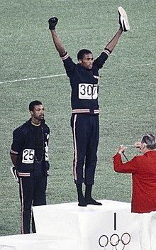 John Carlos (left) and Tommie Smith (center) wearing black gloves, black socks, and no shoes at the 200 m award ceremony of the 1968 Olympics John Carlos, Tommie Smith 1968.jpg
