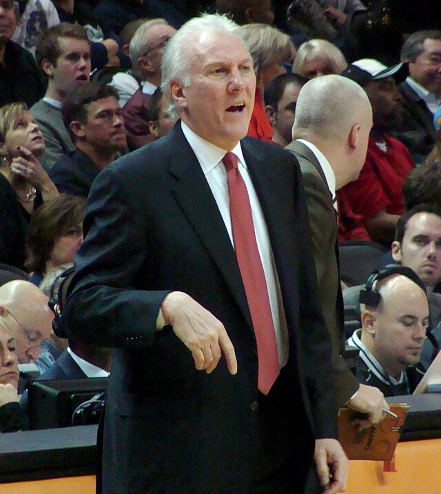 A man, wearing a black suit, white shirt and red tie, is standing in front of the spectators.