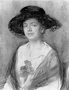 A portrait of a white woman wearing a large dark hat and a white dress, with a translucent dark shawl and flowers pinned to her bodice.