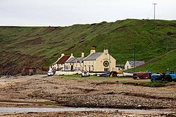 A colection of small buildings in front of a cliff, with a pebble beach in front