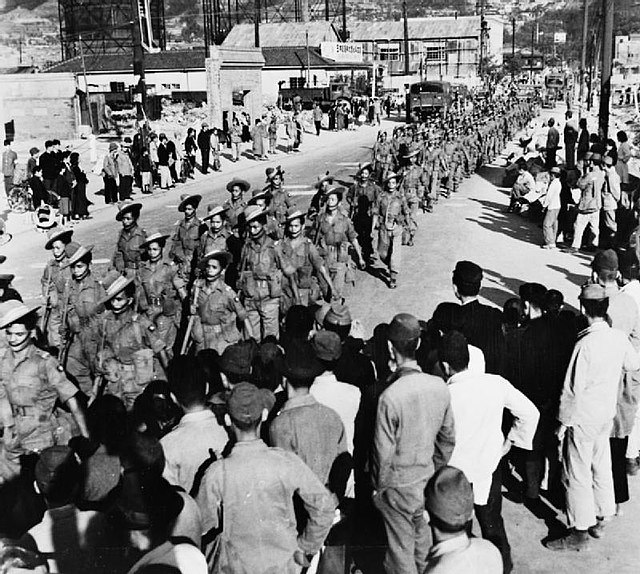 Photo noir et blanc de militaires défilant dans une rue pendant que des civils regardent sur le bas-côté, dans une ville partiellement en ruines.