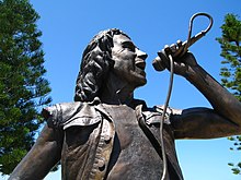 Scott's life-sized statue shown in upper body shot. He sings into a hand held microphone, his chest partly exposed beneath a vest.