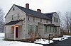 A two-story gray wooden house with light green shutters seen in winter, with snow on the ground. A long wing with two brick chimneys projects towards the left from a higher rear section. At lower right is a black mailbox with "5Ave" on it next to a clear paved driveway.