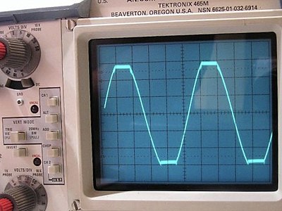 The picture shows an oscilloscope screen of an amplifier "clipping."  The amplifier should be outputting a clean sine wave with rounded tops and bottoms, but instead they are cut off flat, or "clipped", as if they had been cut with a pair of scissors or shears.