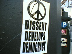 Photograph of a sticker on a public installation that states "DISSENT DEVELOPS DEMOCRACY" in all-caps with a peace symbol above it