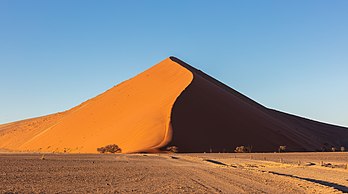 Enorme duna a 39 km do principal ponto de acesso ao parque nacional Namib-Naukluft na estrada para Sossusvlei (às vezes escrito) Sossus Vlei, na Namíbia. Sossusvlei (nome de origem mista, que significa aproximadamente "pântano sem saída") é uma bacia endorreica salina, para o efêmero rio Tsauchab, cercada por altas dunas de cor rosa a laranja vívida, uma indicação de uma alta concentração de ferro na areia e consequentes processos de oxidação. As dunas mais antigas são as de cor avermelhada mais intensa. Essas dunas estão entre as mais altas do mundo; muitas delas estão acima de 200 metros, sendo a mais alta aquela apelidada de Big Daddy, com cerca de 325 metros de altura. O nome "Sossusvlei" é frequentemente usado com um significado extenso para se referir à área circundante (incluindo outros vleis vizinhos, como o Dead Vlei e outras dunas altas), que é uma das principais atrações turísticas do país. (definição 8 112 × 4 529)