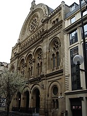 Seitliche Farbfotografie eines braunen Gebäudes mit Rundbogenfenstern und Steinrosetten. Der Eingang besteht aus drei Rundbögen. Das Runddach hat eine große Steinrosette.
