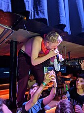 Photo of Joost Klein standing on a bar, not wearing a short, holding a microphone