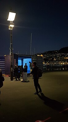 blue light emits from a shipping container with people walking around at night time