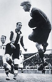 Photographie en noir et blanc d'un gardien de football, de dos, captant le ballon, devant des attaquants adverses.