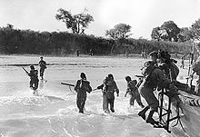 men climbing down side of small boat into the surf on to a sandy beech with trees in the background