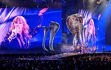 Swift performing onstage with a graphic of a striking cobra and a close-up shot of her behind her.