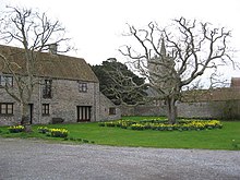 Photo of large, two-storey stone building with gabled roof and surrounding stone wall. The house has a gravel driveway and large grassy space in front with yellow daffodils and two bare trees. A village church tower is located in the distance, beyond the wall.