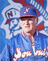 A man in glasses wearing a blue satin jacket with "Sounds" written on the front in white and red trim with a blue cap with a white "N" on the front