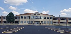 A photo of the front of a two story building with a creme colored facade and red brick columns every few feet. The building has lots of windows. Blue and grey letters in some of the windows spell the words "Cascade Cadets". Benches and garbage cans flank the doors. A large tree is visible to the viewer's left.